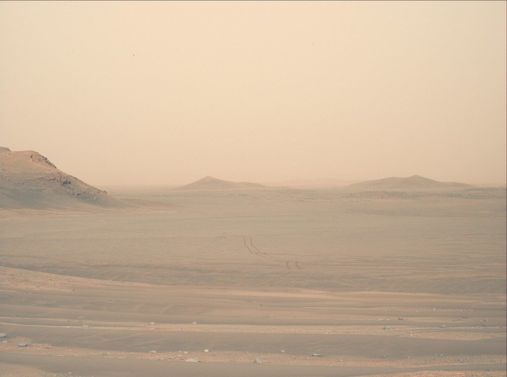 Faint rover wheel tracks curve across a dusty plain on Mars toward low hills on the horizon.