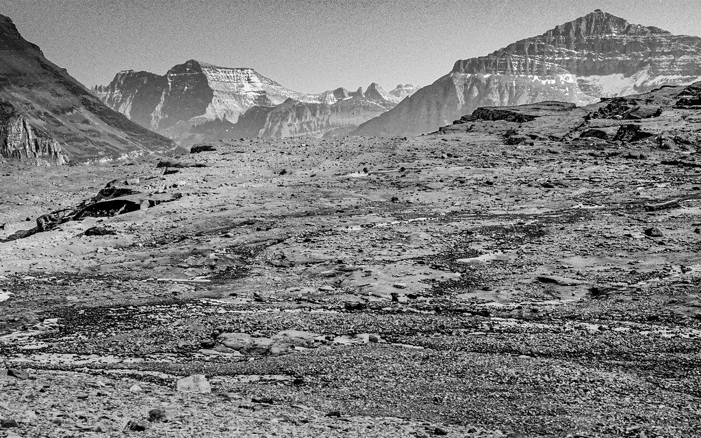 A black and white photograph of bare rock and rubble filling a mountain basin where a glacier once stood, with the same ridges on the skyline as in the earlier view.