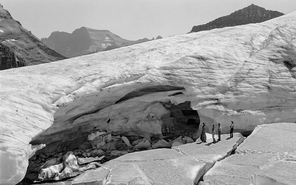A black and white photograph of four people standing beside the snout of a glacier, where meltwater has hollowed a cave in the ice, with mountain ridges behind.