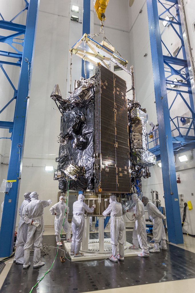 A tall satellite wrapped in black and silver insulation stands between blue support towers while technicians in white coveralls work around its base.