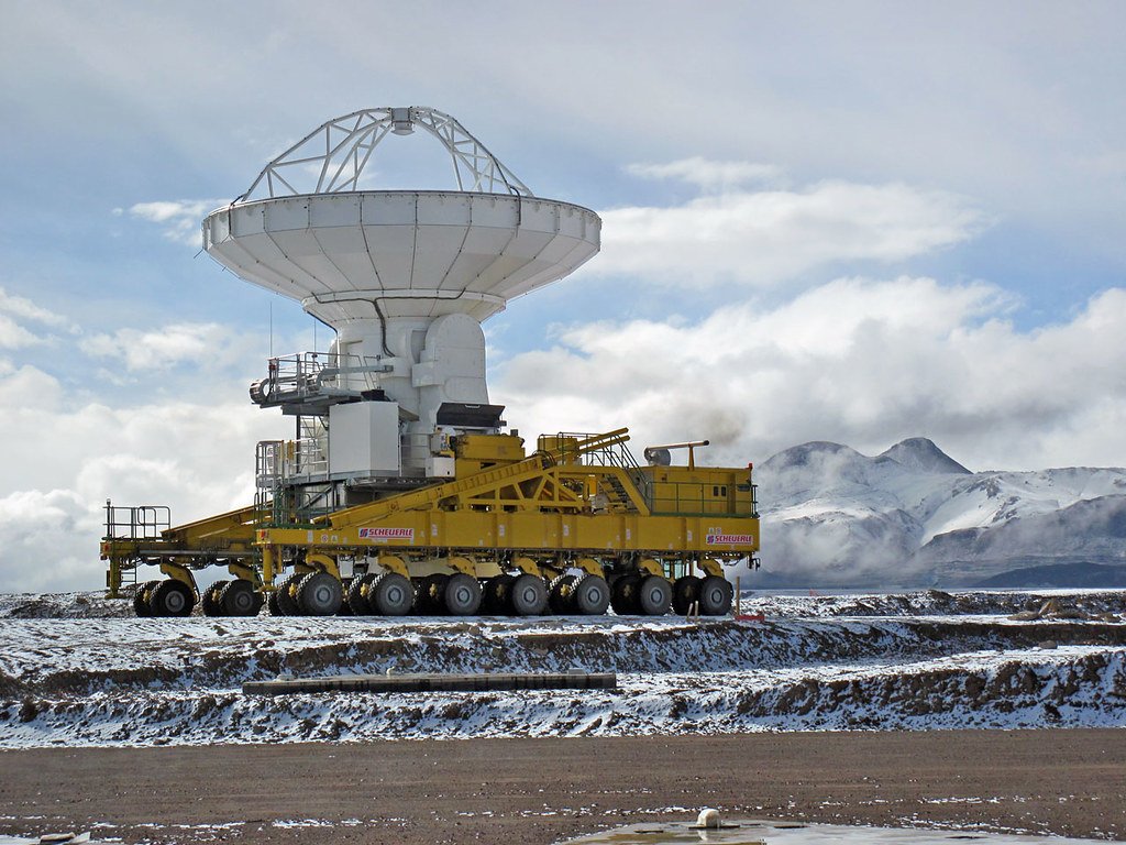 A yellow multi-wheeled transporter carrying a white radio antenna over snow-dusted ground, with snowy peaks behind.