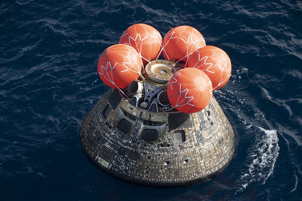 An Orion capsule floats in dark blue ocean water with five orange flotation bags inflated around its upper rim.