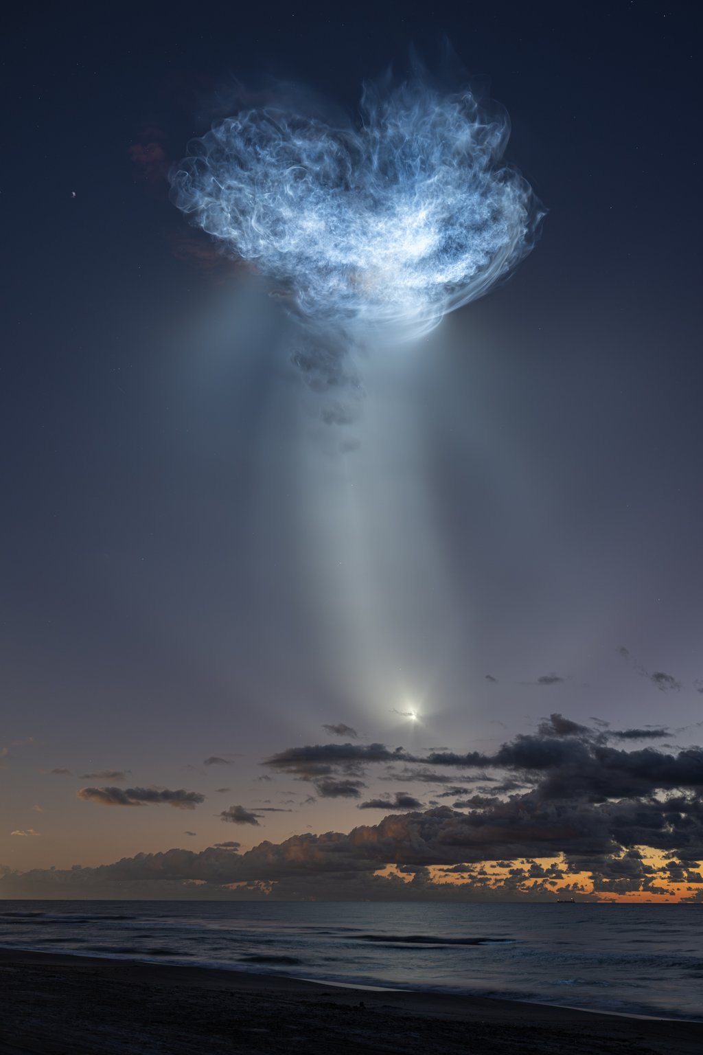 A tall, sunlit rocket exhaust plume stands like a column in a twilight sky above a line of dark clouds and an orange horizon, with the ocean and a dark beach below.