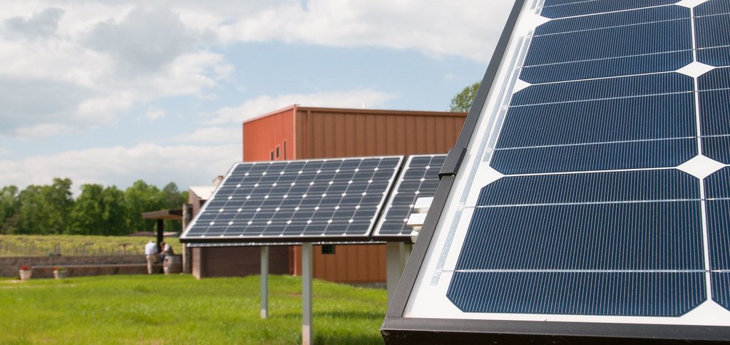 A ground-mounted solar panel in close-up with two more panels on frames behind it, on grass beside a winery building.
