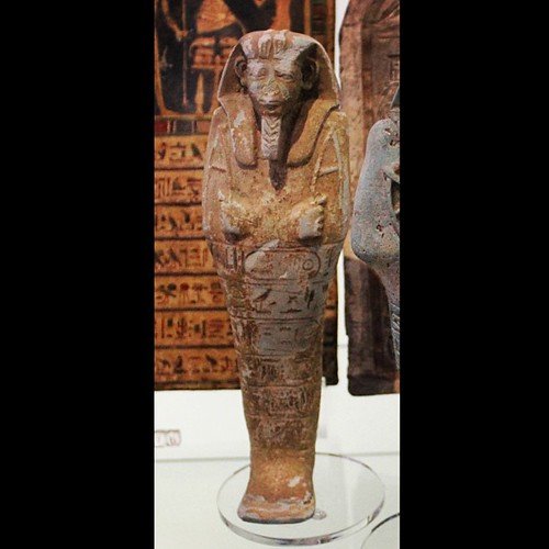 A small glazed faience figure of a mummiform king covered in carved hieroglyphs, standing in a museum display case.