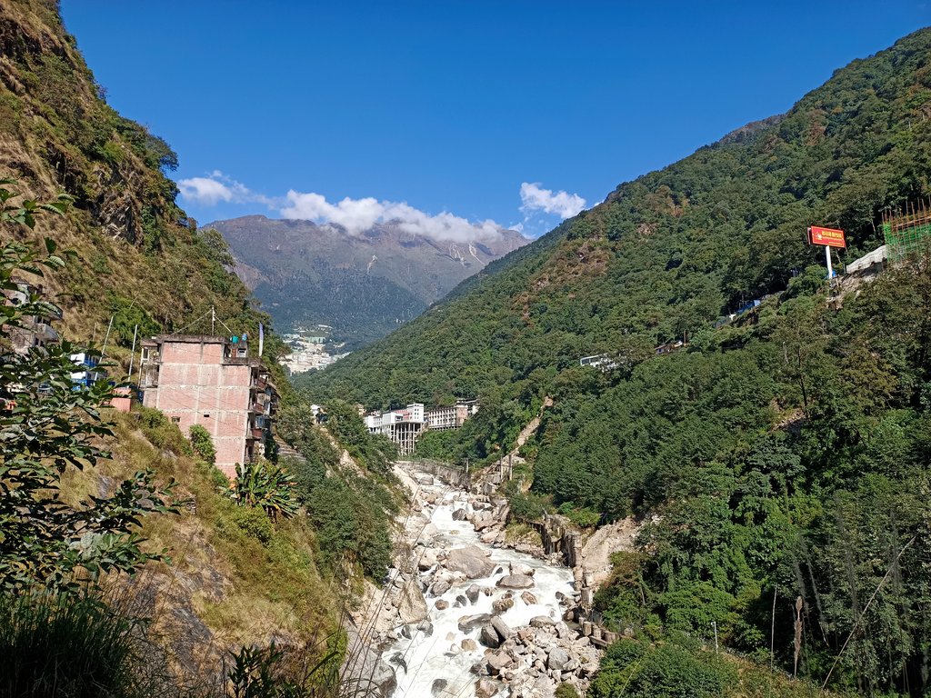 A narrow forested gorge at the Nepal-China border, a boulder-filled river running through the bottom and concrete buildings standing on the slope above it.