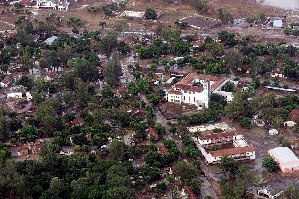 Aerial view of a town of tin-roofed houses and trees, with standing water and mud along its streets.