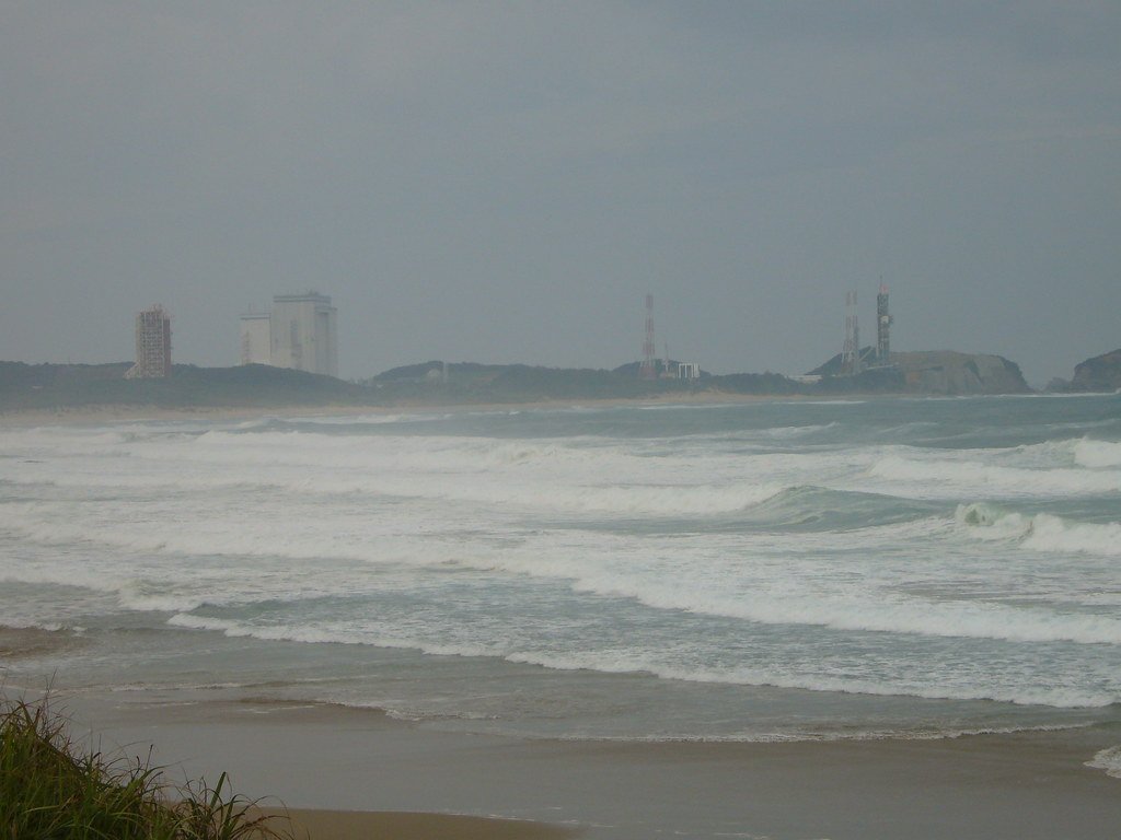 Two launch service towers and white assembly buildings on a low green headland, seen from a beach across a stretch of breaking surf.