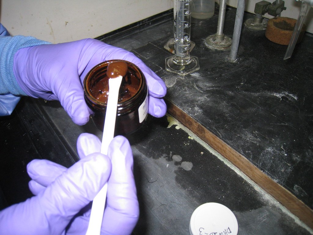 Gloved hands hold a sample jar of thick brown emulsified oil on a laboratory bench, with a graduated cylinder behind.