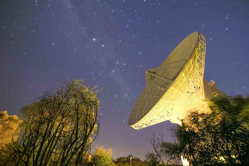 A large dish antenna lit from below against a star-filled night sky, framed by trees.