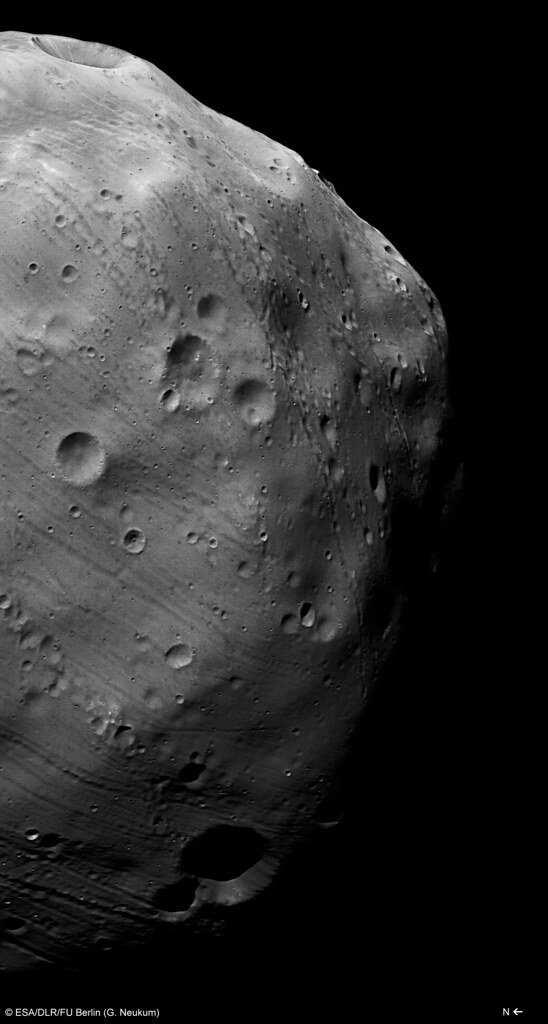 A close pass over the cratered surface of a small irregular moon, seen edge-on with its limb against black space.