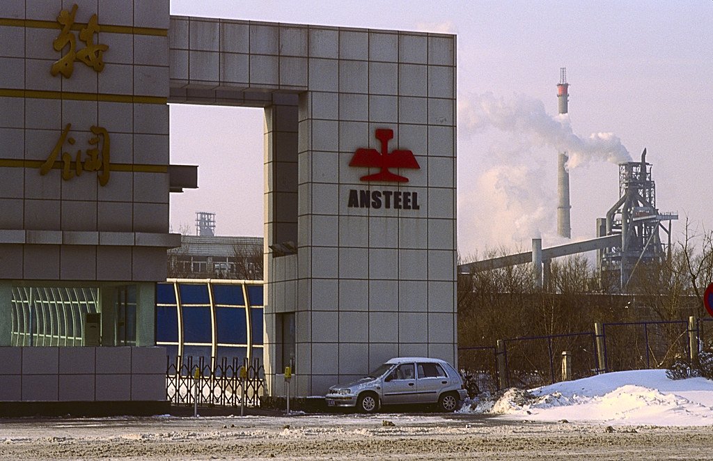 The entrance gate of a Chinese steel complex in winter, with a blast furnace and a smoking stack standing behind it.