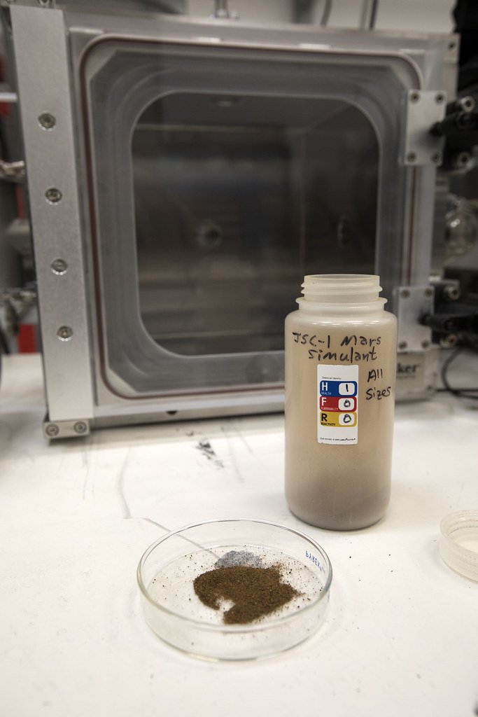 A jar labeled Mars simulant by hand and a petri dish of brown powder on a bench in front of a laboratory chamber.