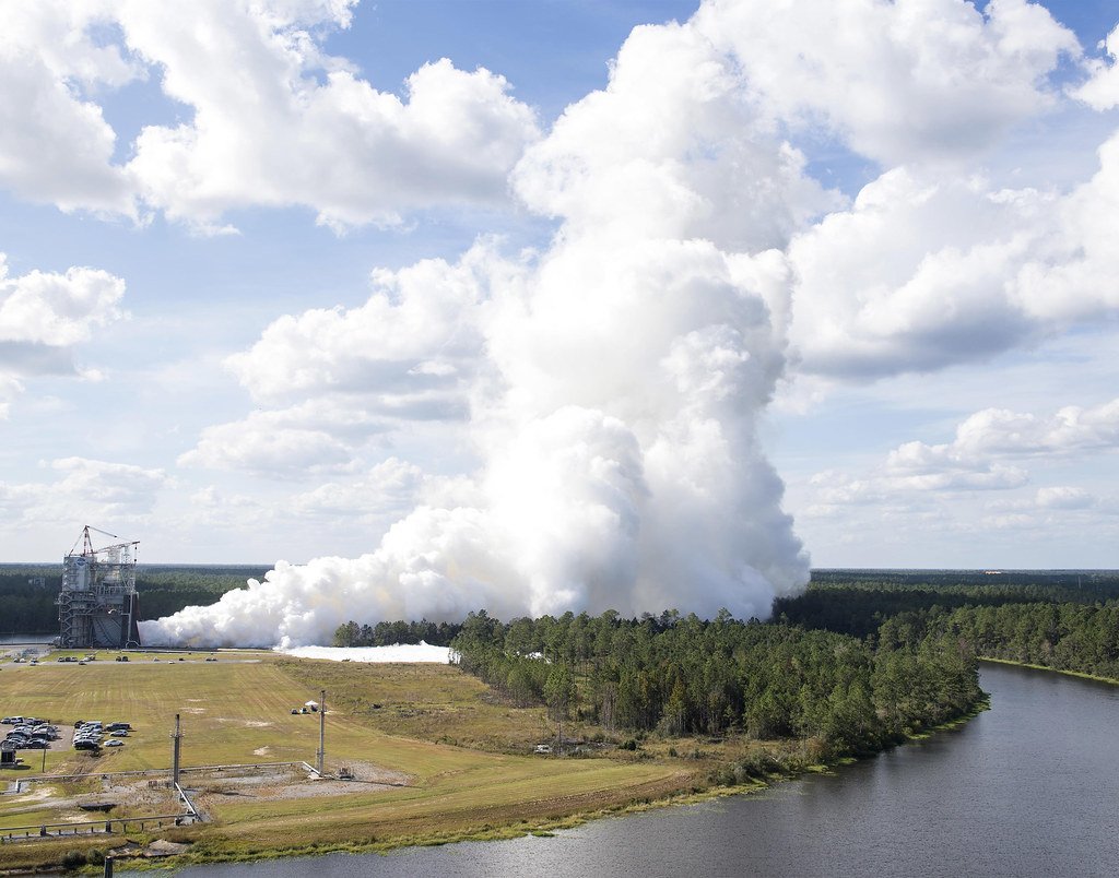 An aerial view of a white steam column rising high above pine forest, with a test stand at the left edge and a waterway at the right.