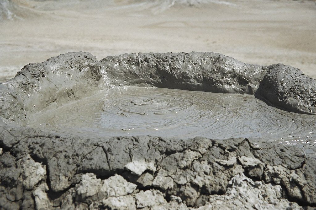 A low crater of dried gray mud on cracked ground, its pool rippled in rings from the center.