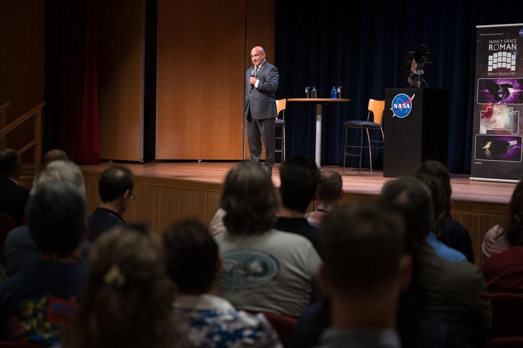 A speaker with a microphone on an auditorium stage facing a seated audience, with a Nancy Grace Roman Space Telescope mission banner standing at the right of the stage.