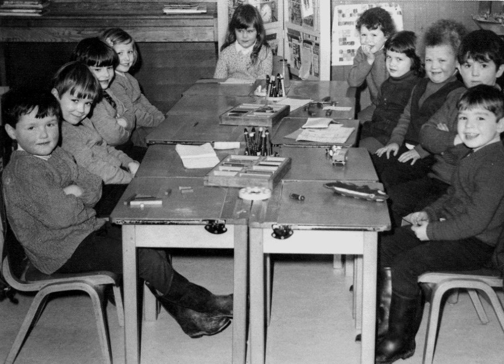 A black-and-white photograph of young schoolchildren seated around joined wooden desks in a classroom, with pencil trays, papers and model boats in front of them.