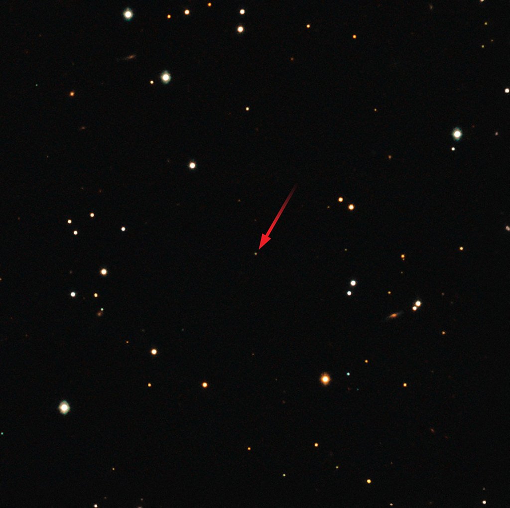 A dark star field photographed from the ground, with a red arrow pointing at a faint orange point of light.