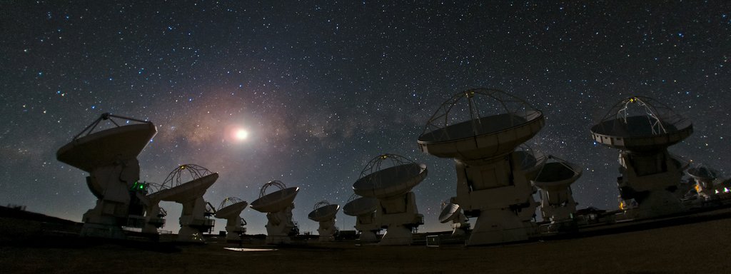 A row of large dish antennas on a high desert plateau at night, tilted toward a sky crowded with stars.
