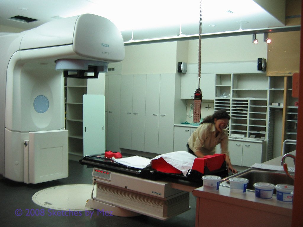 A white linear accelerator with its gantry arm extended over a treatment couch, in a clinic room where a staff member is working at a counter.