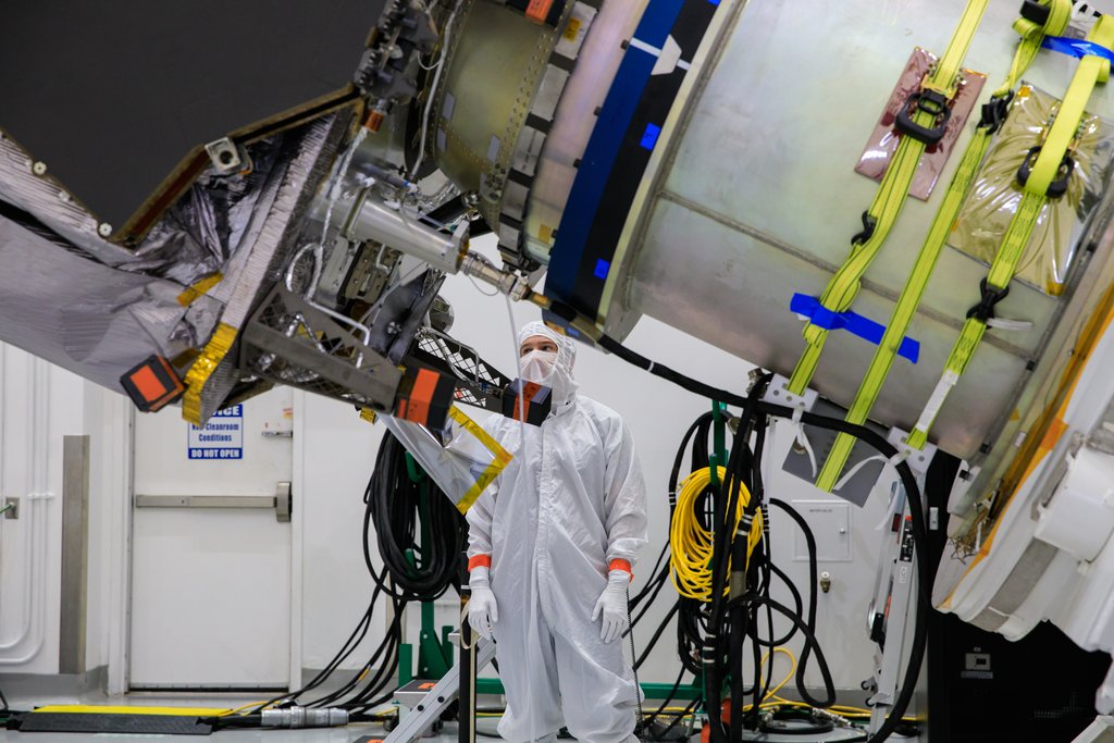 A technician in a white clean-room suit stands beneath the Lucy spacecraft, held horizontally in a rotation fixture among cables and lifting straps.