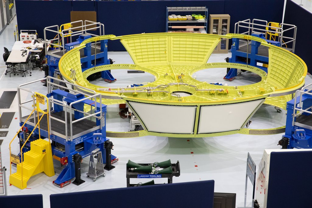 A large yellow ring-shaped rocket structure held in blue and silver assembly tooling on a factory floor.