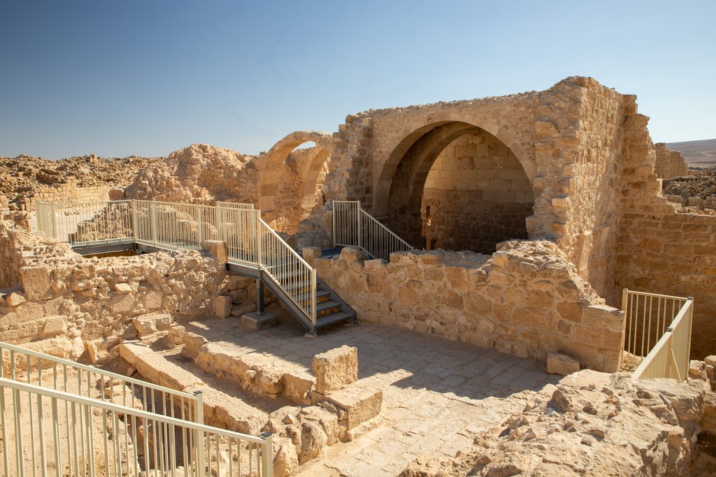 A vaulted stone building standing among low ruined walls and paved courtyards, with modern metal walkways and handrails threading between them and open desert on the horizon.