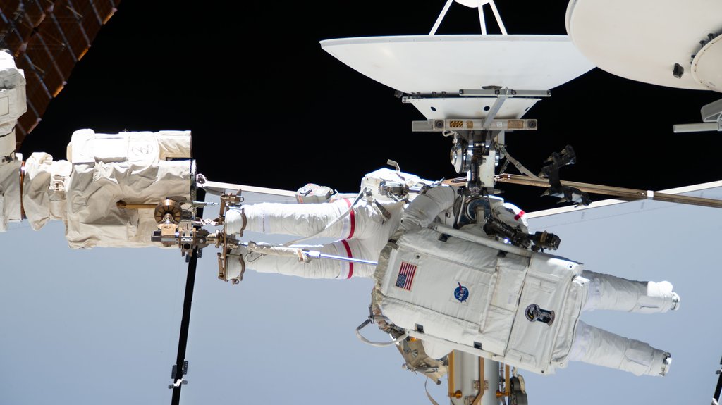 A suited astronaut works amid antennas and handrails on the outside of the space station, with Earth below the horizon at lower left.