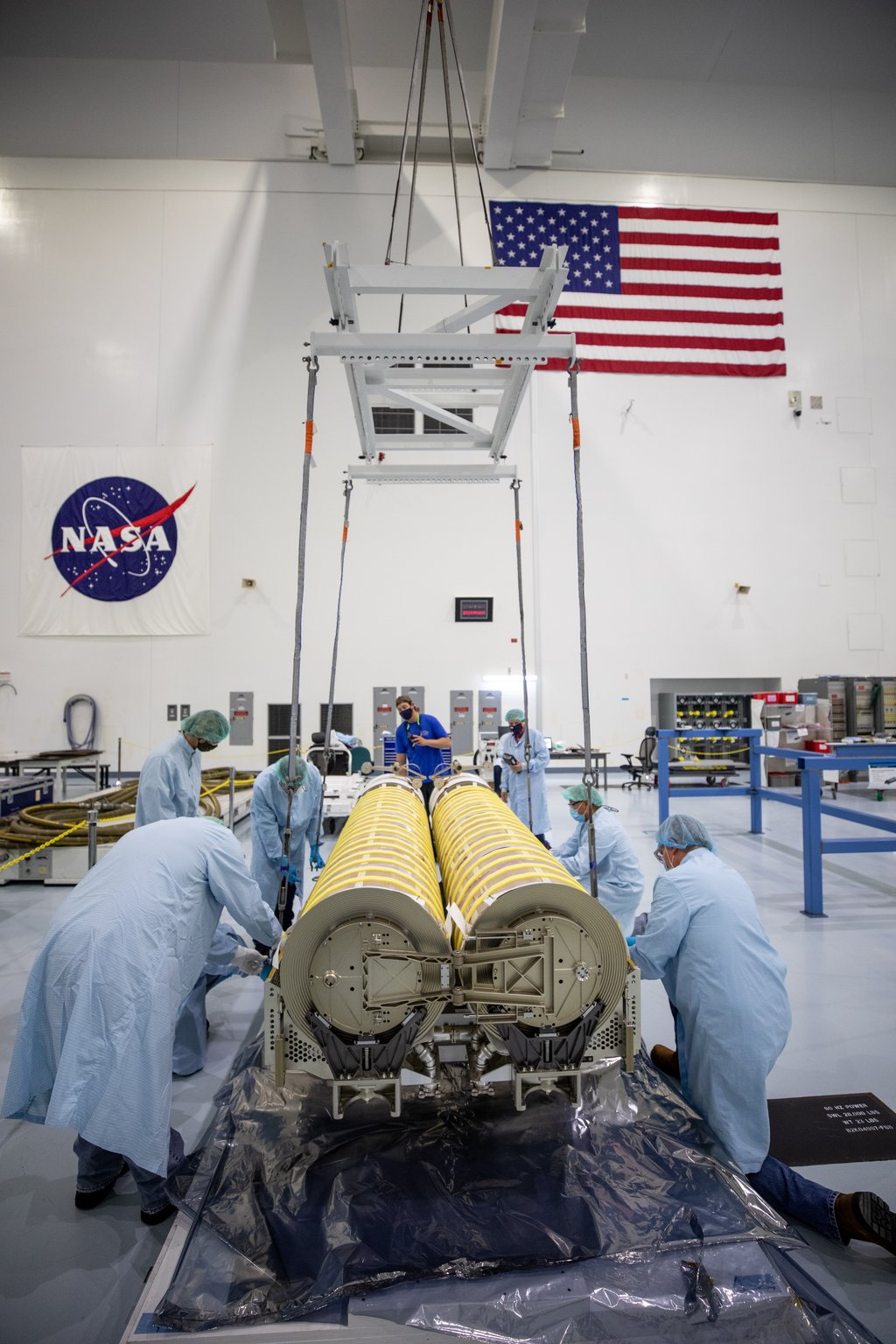 Workers in blue coveralls guide two rolled-up solar array canisters onto a pallet inside a clean high bay, with a NASA banner and a US flag on the wall behind them.