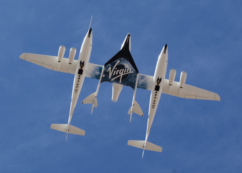 Seen from the ground, a twin-fuselage aircraft flies overhead with a small spaceplane carried between its fuselages against a blue sky.