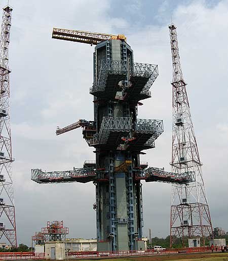 An empty launch pad with a tall gray service tower, folded access platforms and two red and white lightning towers standing on either side.