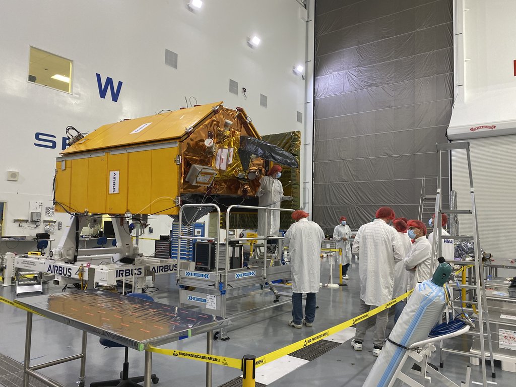The gold and white Sentinel-6 Michael Freilich satellite stands on a support stand in a high-bay cleanroom with technicians working around it.