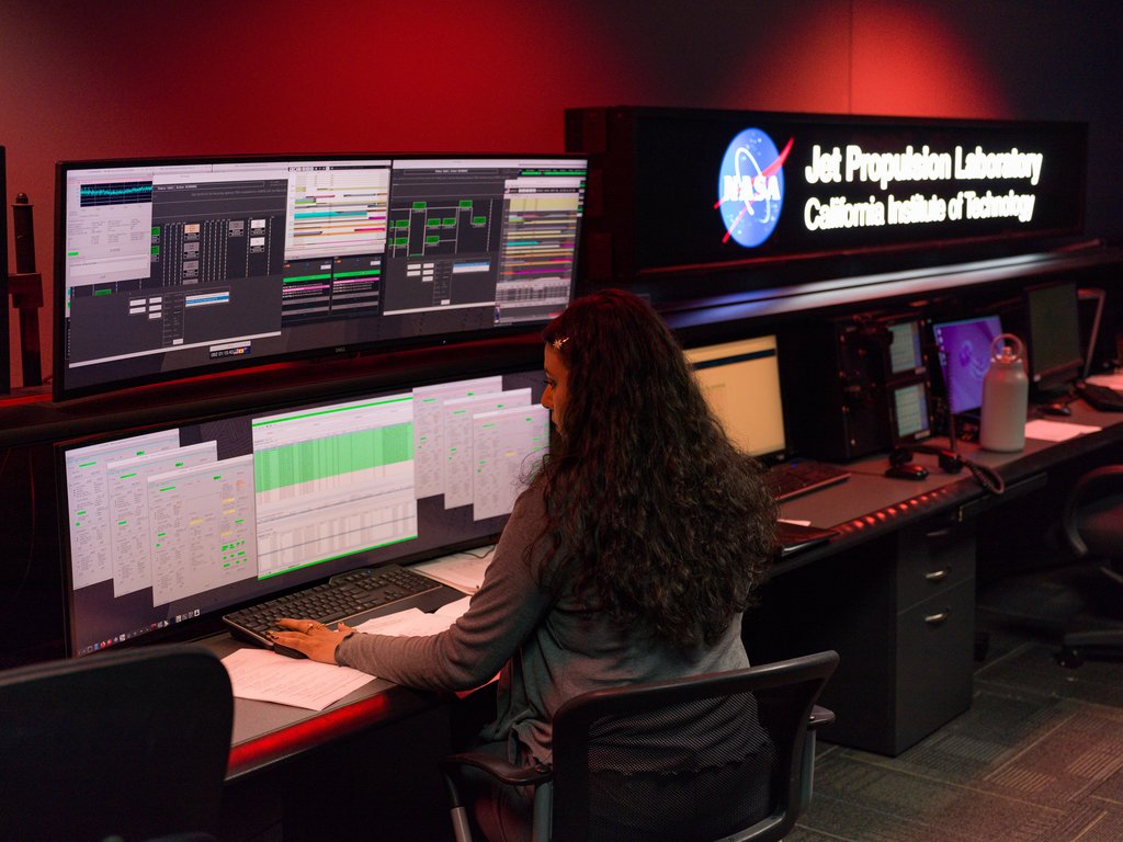 An operator seated at a console of screens showing tracking schedules and link status in a darkened control room.