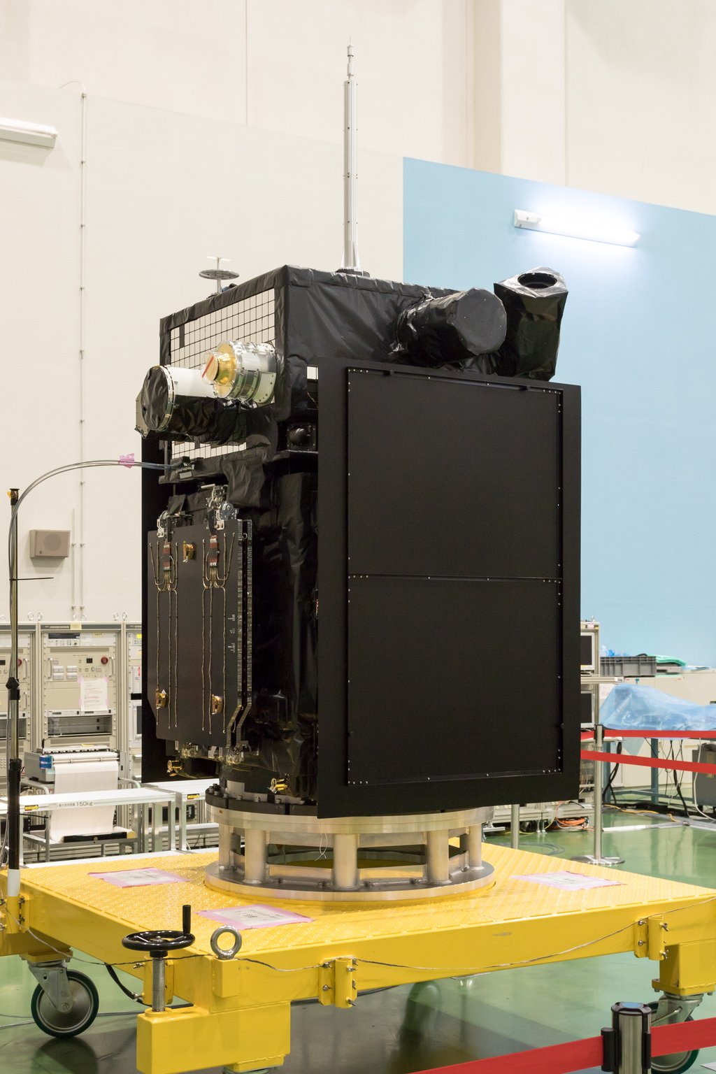 The boxy Arase satellite, wrapped in black thermal blanketing, standing on a yellow handling cart in an assembly hall.